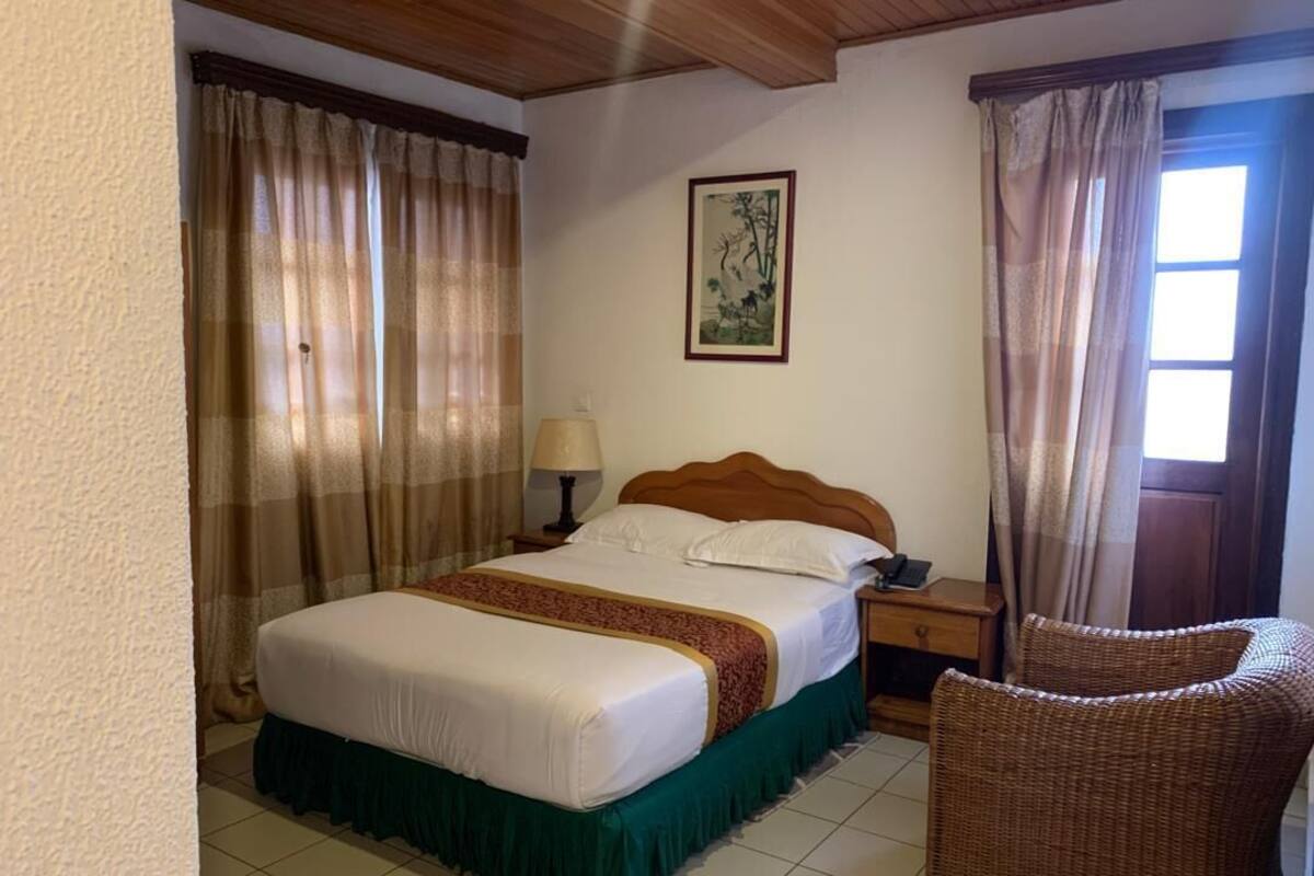 Een hotelkamer met een bed, een nachtkastje, een lamp, een rieten stoel en een schilderij aan de muur.
