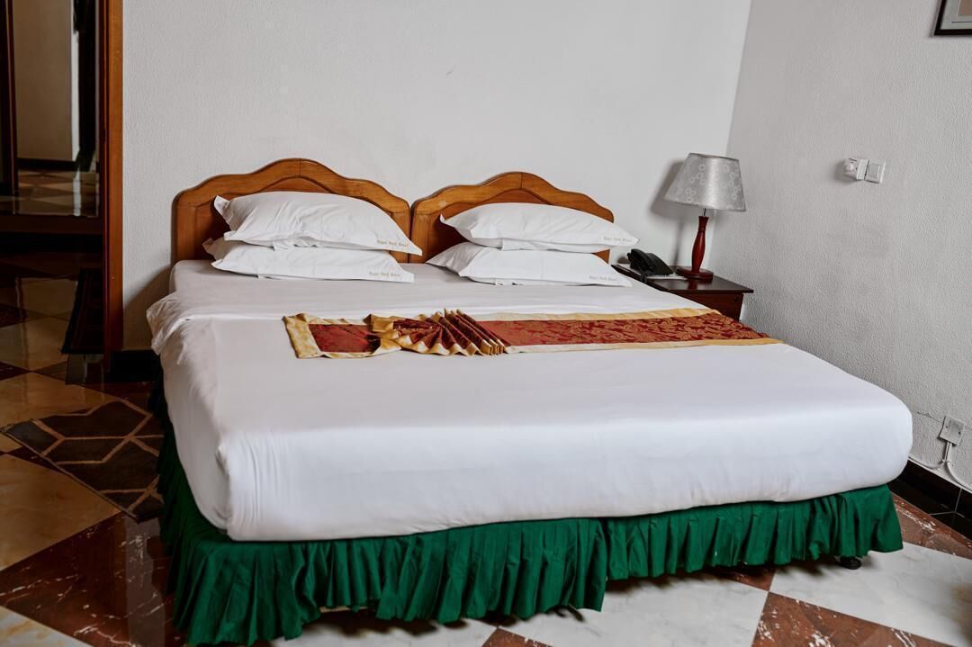 A neatly made bed with white linens and a green skirt, a wooden headboard, a bedside table with a lamp, and a patterned rug.
