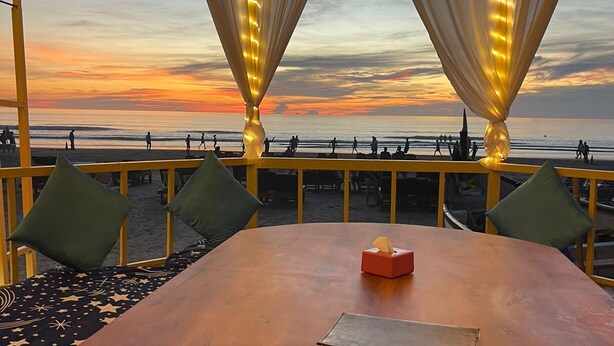 A round wooden table with a tissue box and a book, surrounded by green cushioned seating, overlooking a beach at sunset. - Sydney X cafe and Huts (Arambol)