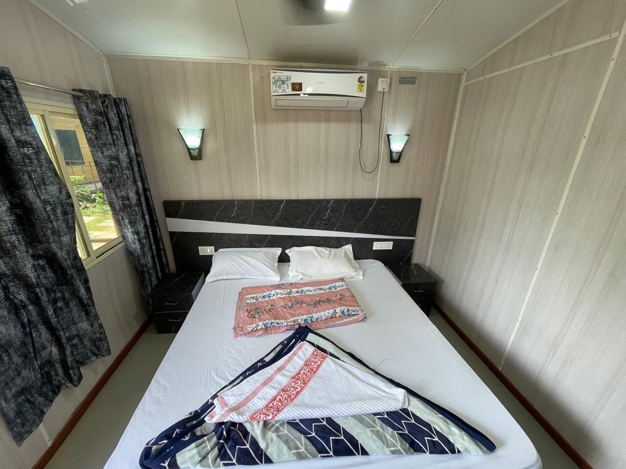 A hotel room with a bed, a wall-mounted air conditioner, a window with curtains, and a wall-mounted light fixture.
