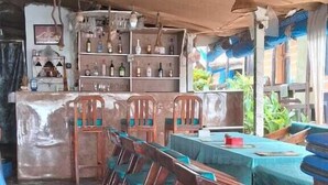 A bar area with wooden chairs and tables, a counter with bottles, and hanging lights. - Sydney X cafe and Huts (Arambol)