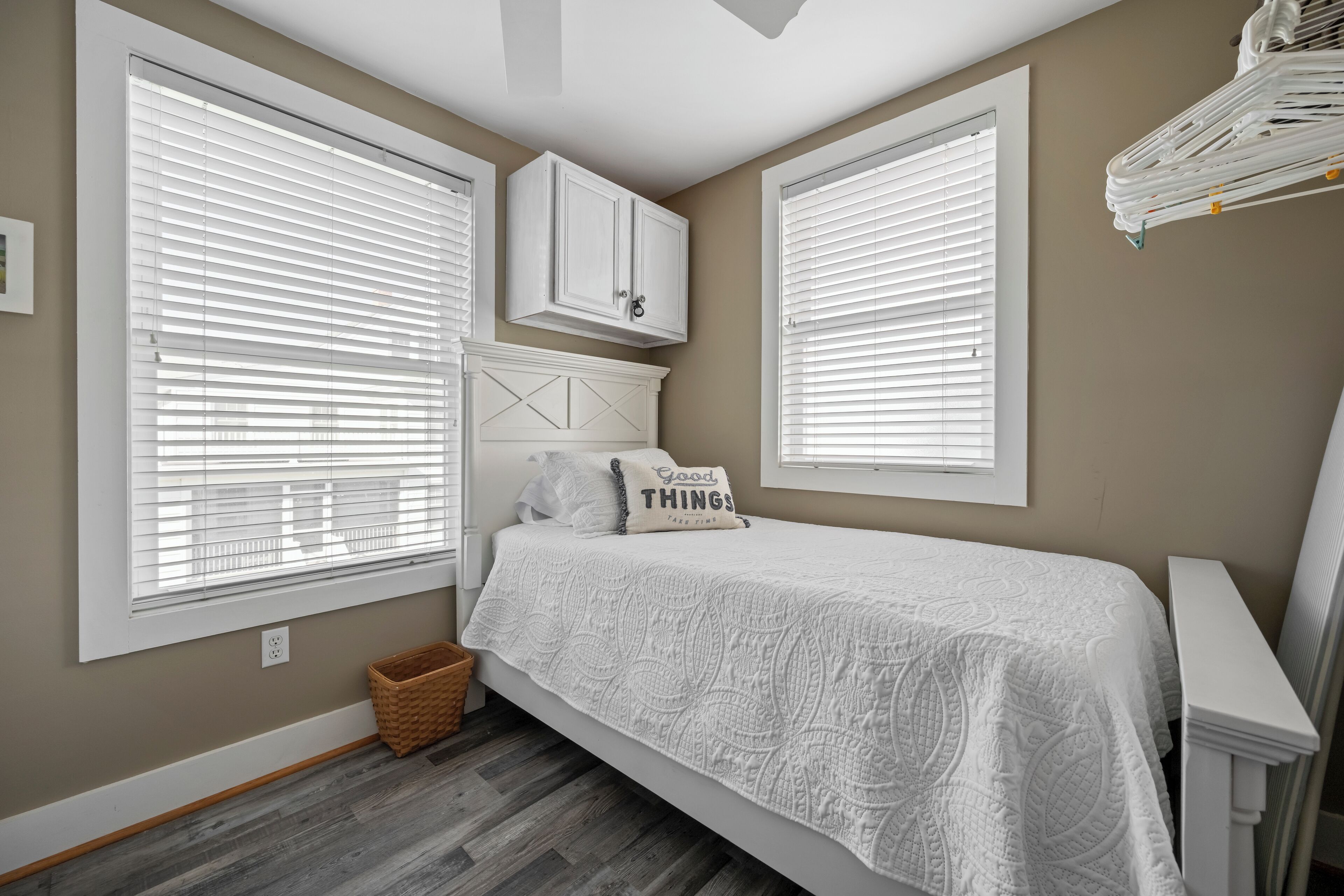 A bedroom with a bed, two windows with blinds, a white headboard, a white cabinet, and a small basket.
