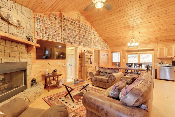 A cozy log cabin interior with a fireplace, a stone wall, a bookshelf, leather furniture, and a kitchen area. - Cottage w/ Hot Tub: 3 Mi to Downtown Blue Ridge! (Blue Ridge)