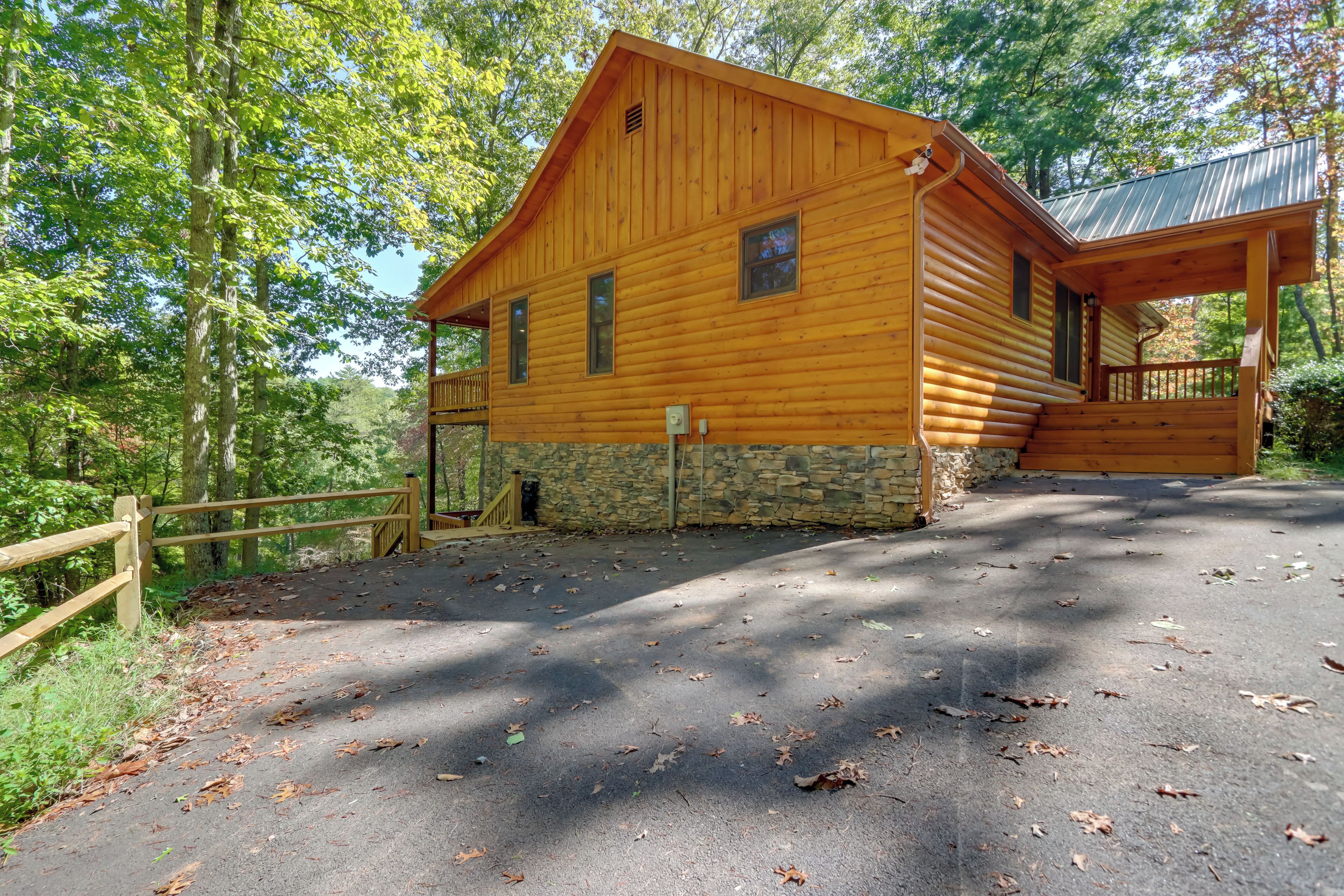 A wooden cabin with a stone foundation, a porch, and a fenced driveway.