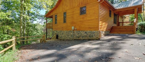 A wooden cabin with a stone foundation, a porch, and a fenced driveway.