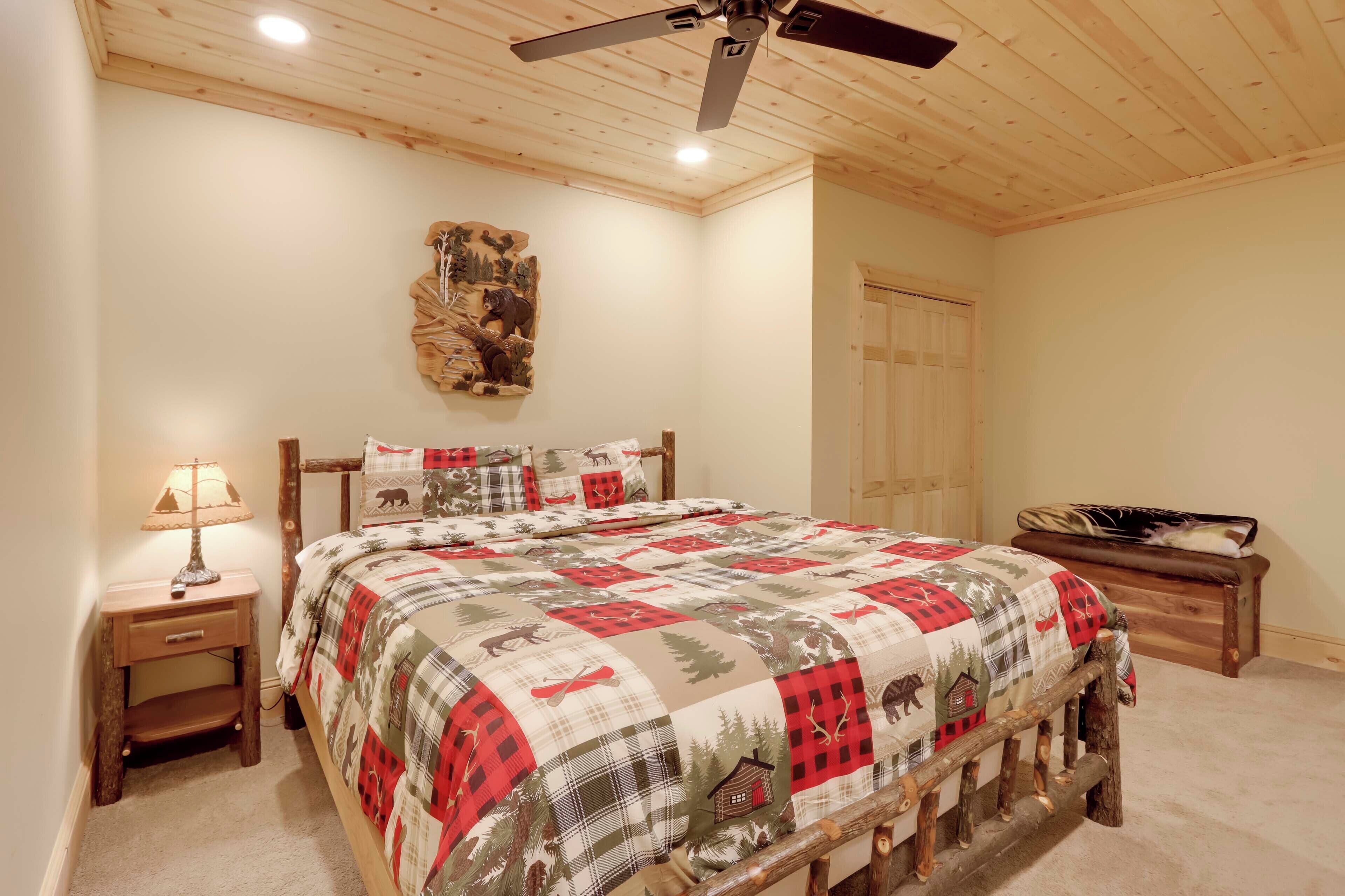 A bedroom with a wooden ceiling, a bed with a plaid bedspread, a nightstand with a lamp, and a wooden bench.
