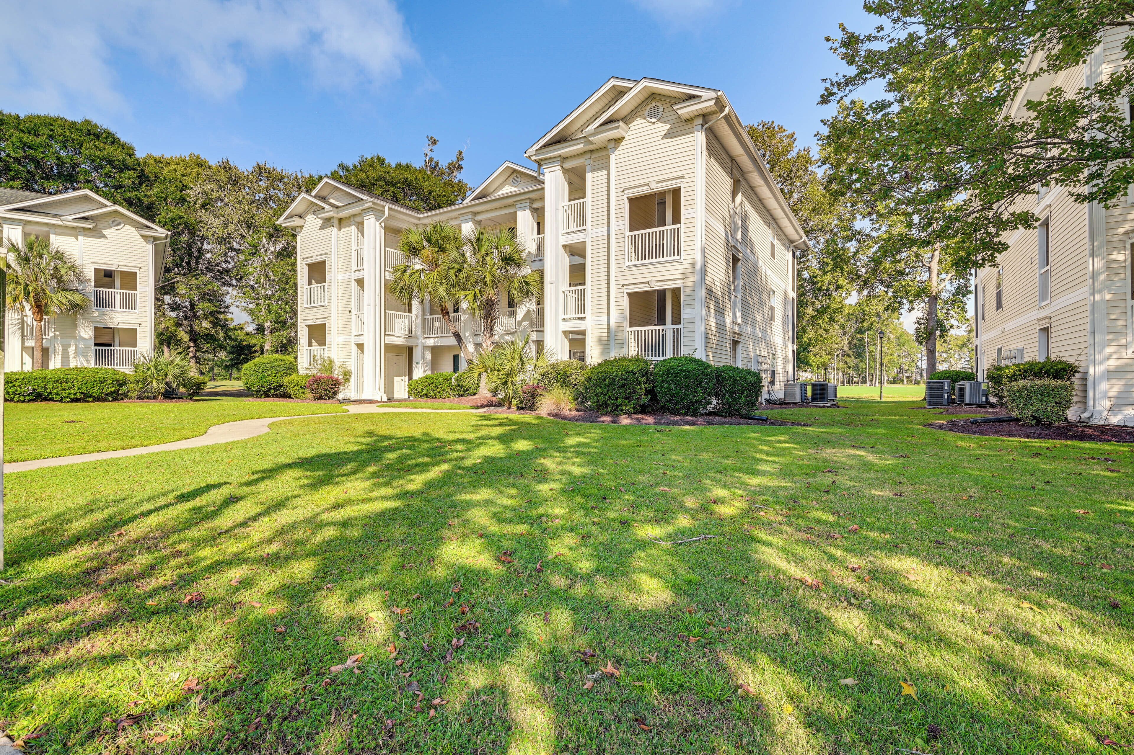 A row of white multi-story buildings with balconies, surrounded by well-maintained lawns and trees.