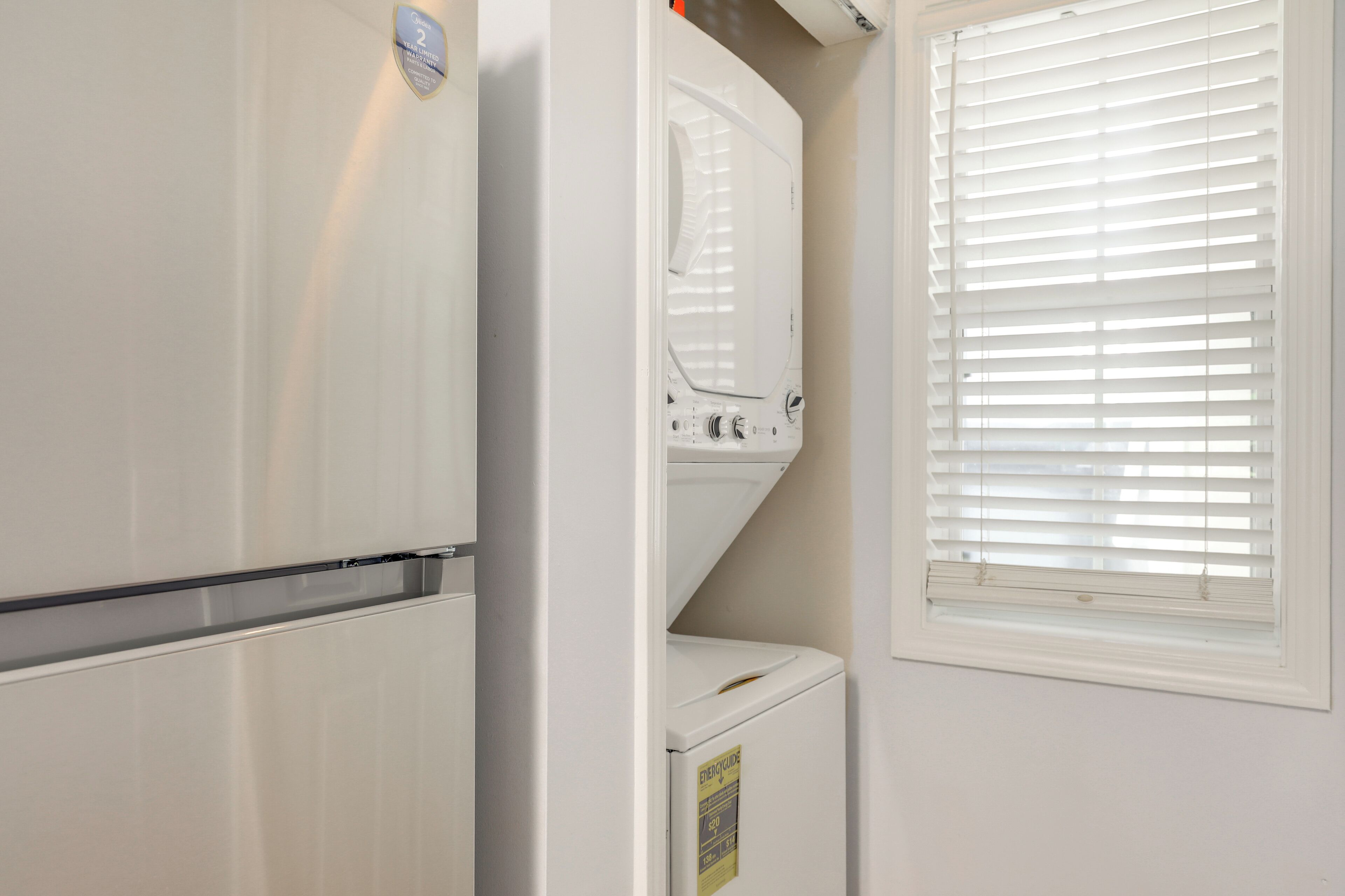 A compact laundry room with a washer and dryer, a window with blinds, and a visible energy efficiency label.