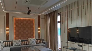 A modern living room with a sectional sofa, ottomans, a coffee table, a flat-screen TV, and a decorative wall panel. - The Taj Mughal Courtyard Homestay (Agra)
