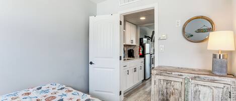 A bedroom with a bed, a white door leading to a kitchen, and a rustic cabinet.
