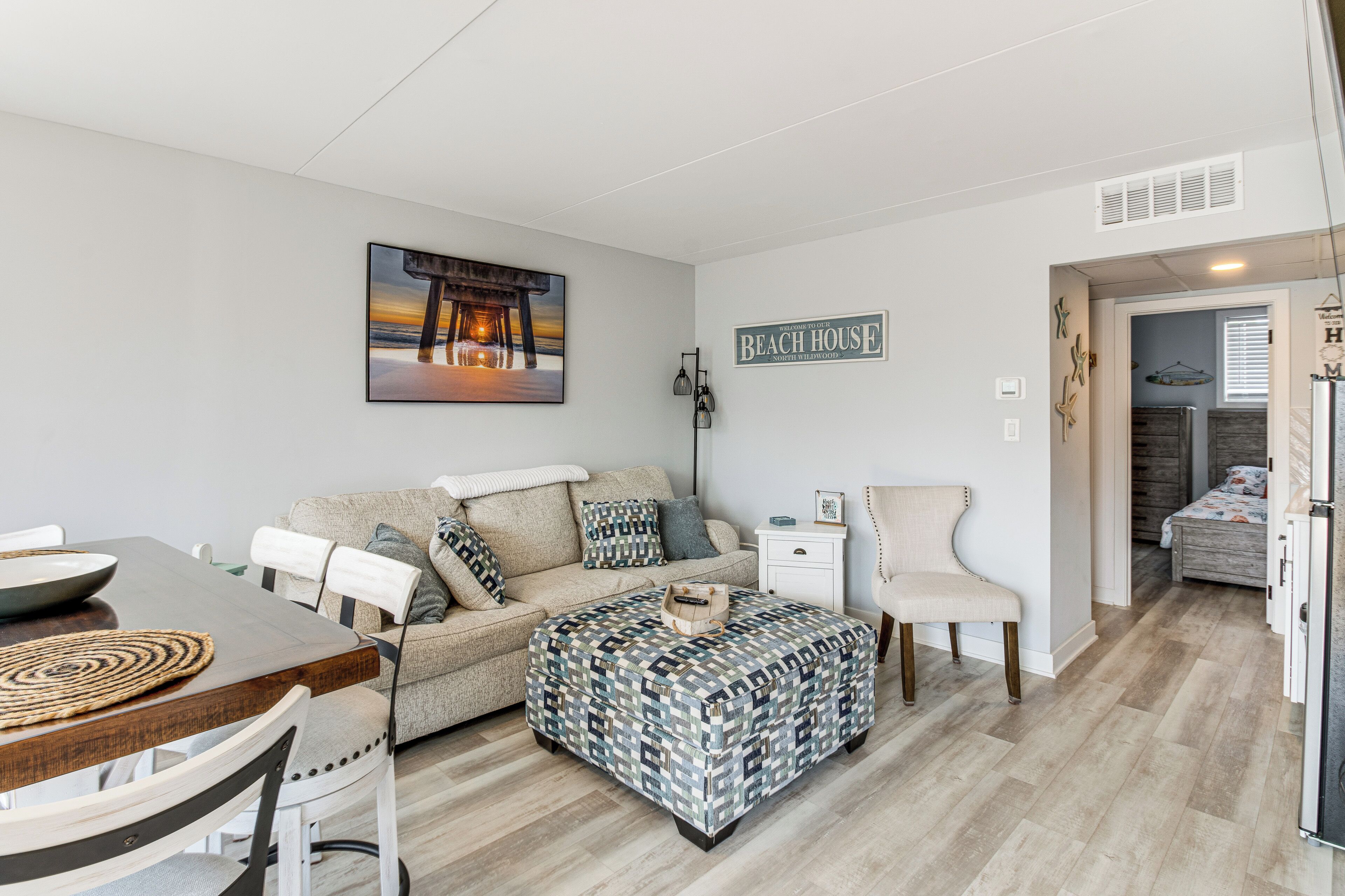 A cozy living room with a sofa, ottoman, and a dining table. There is a painting on the wall and a sign that says "Beach House".