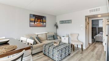 A cozy living room with a sofa, ottoman, and a dining table. There is a painting on the wall and a sign that says "Beach House".