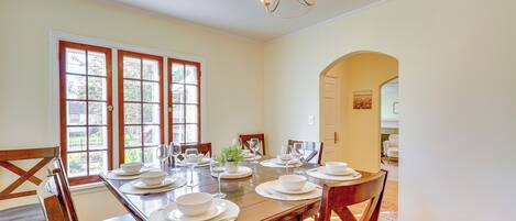Comedor con una mesa de madera dispuesta para seis personas, sillas alrededor, ventanas amplias y una araña.