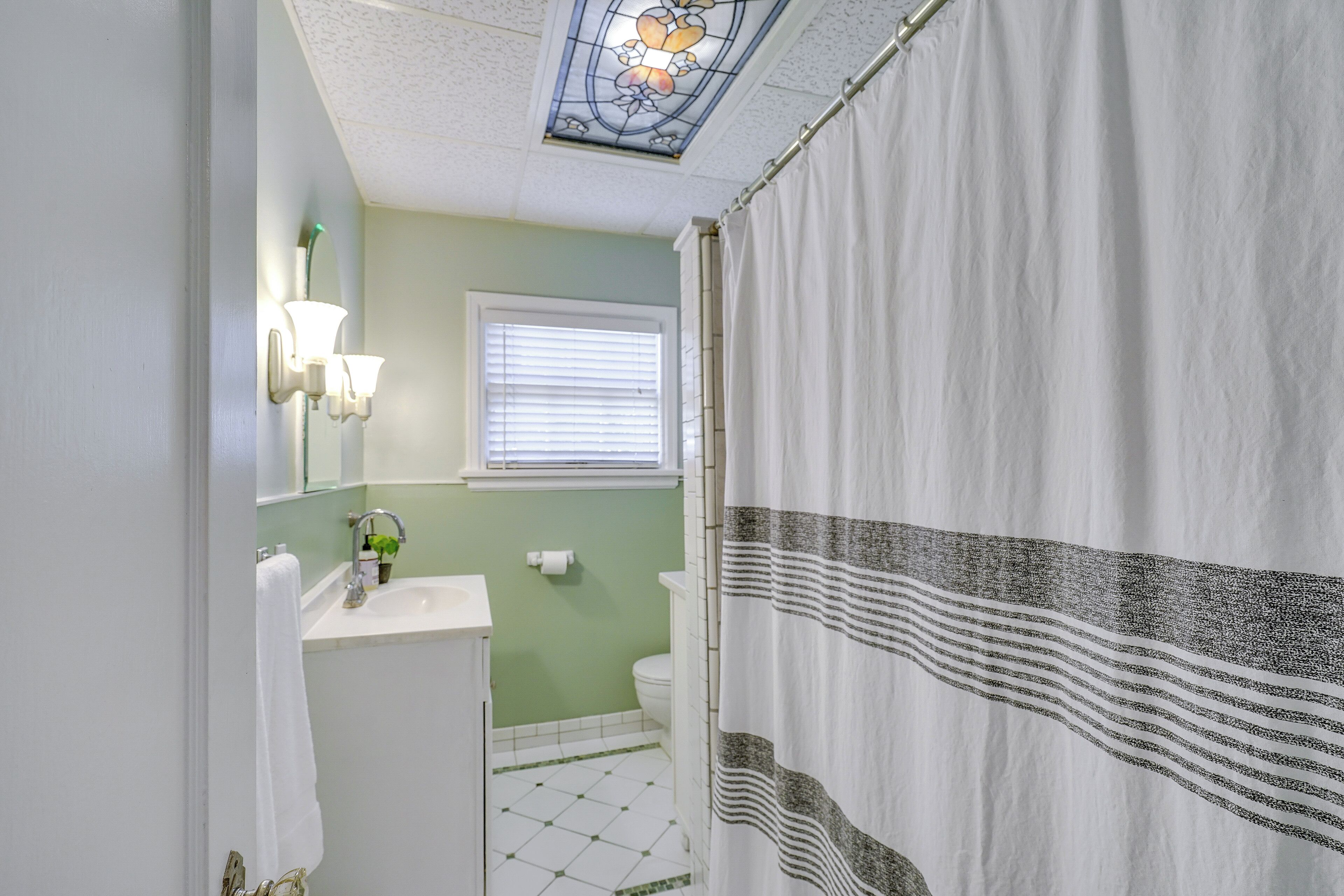 A bathroom with a white sink, a mirror, a window with blinds, and a shower curtain.