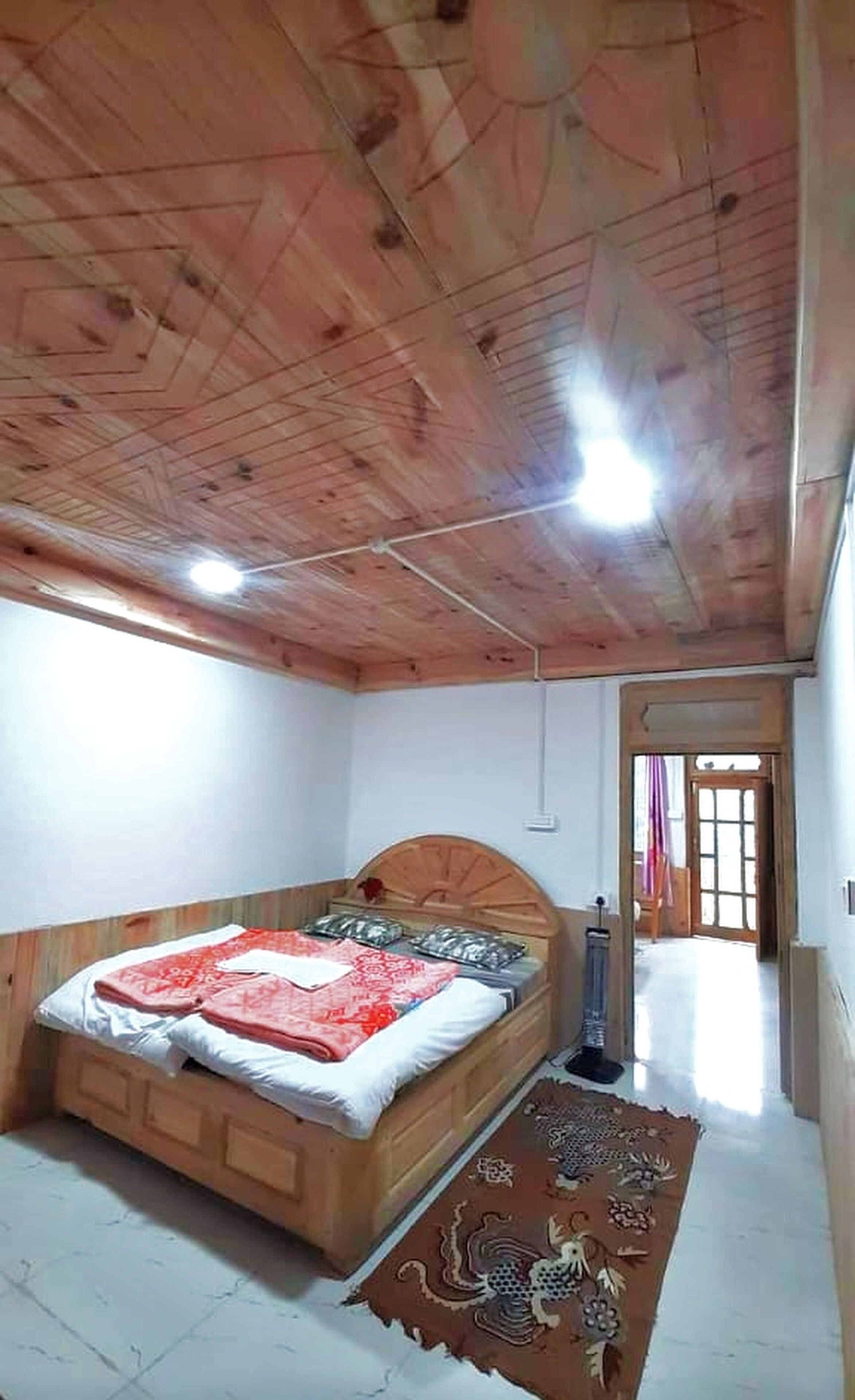 A bedroom with a wooden ceiling, a bed with a red and white bedspread, and a patterned rug on the floor.