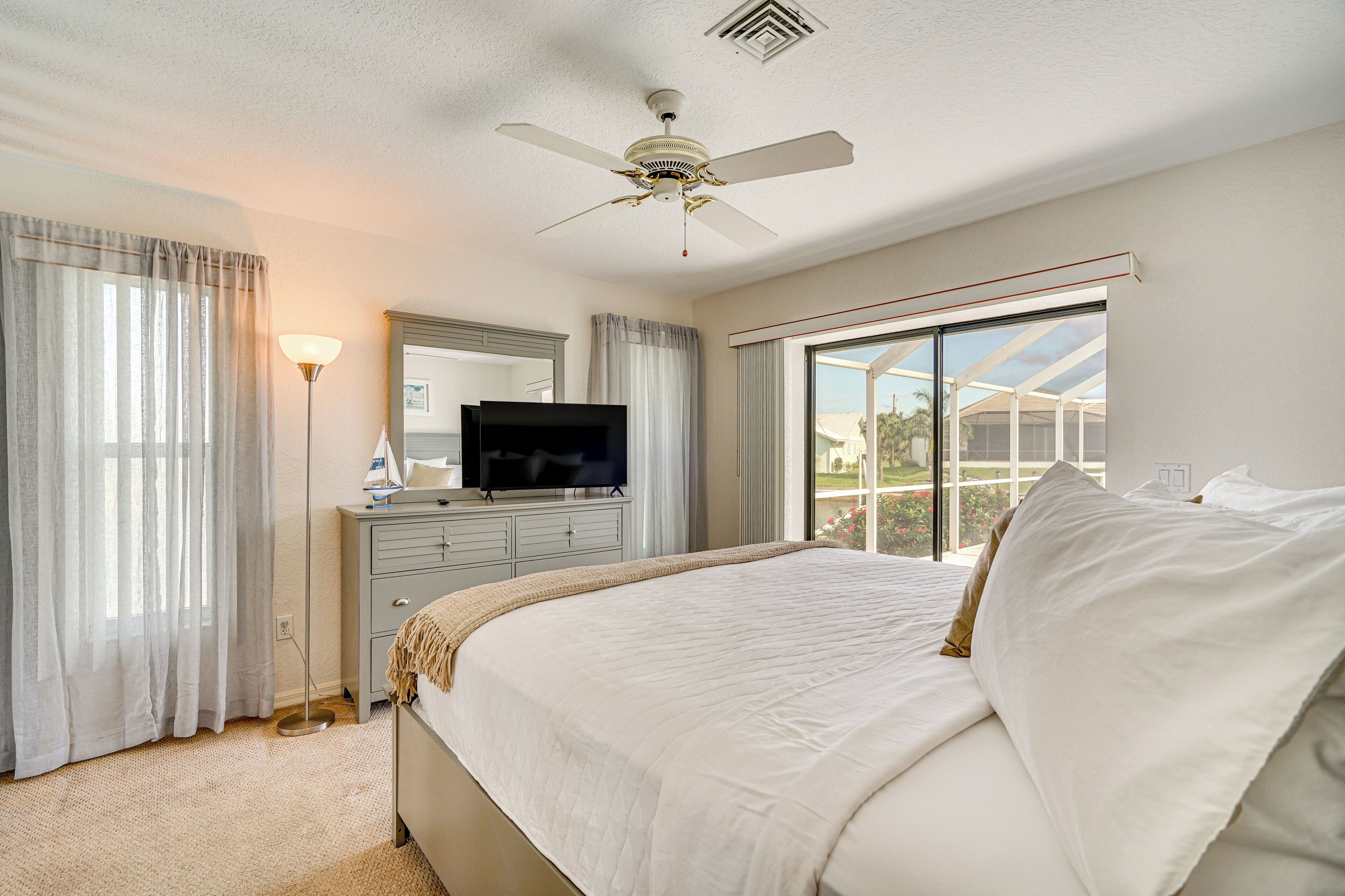 A bedroom with a bed, a ceiling fan, a television, a dresser, and a large window with a view.