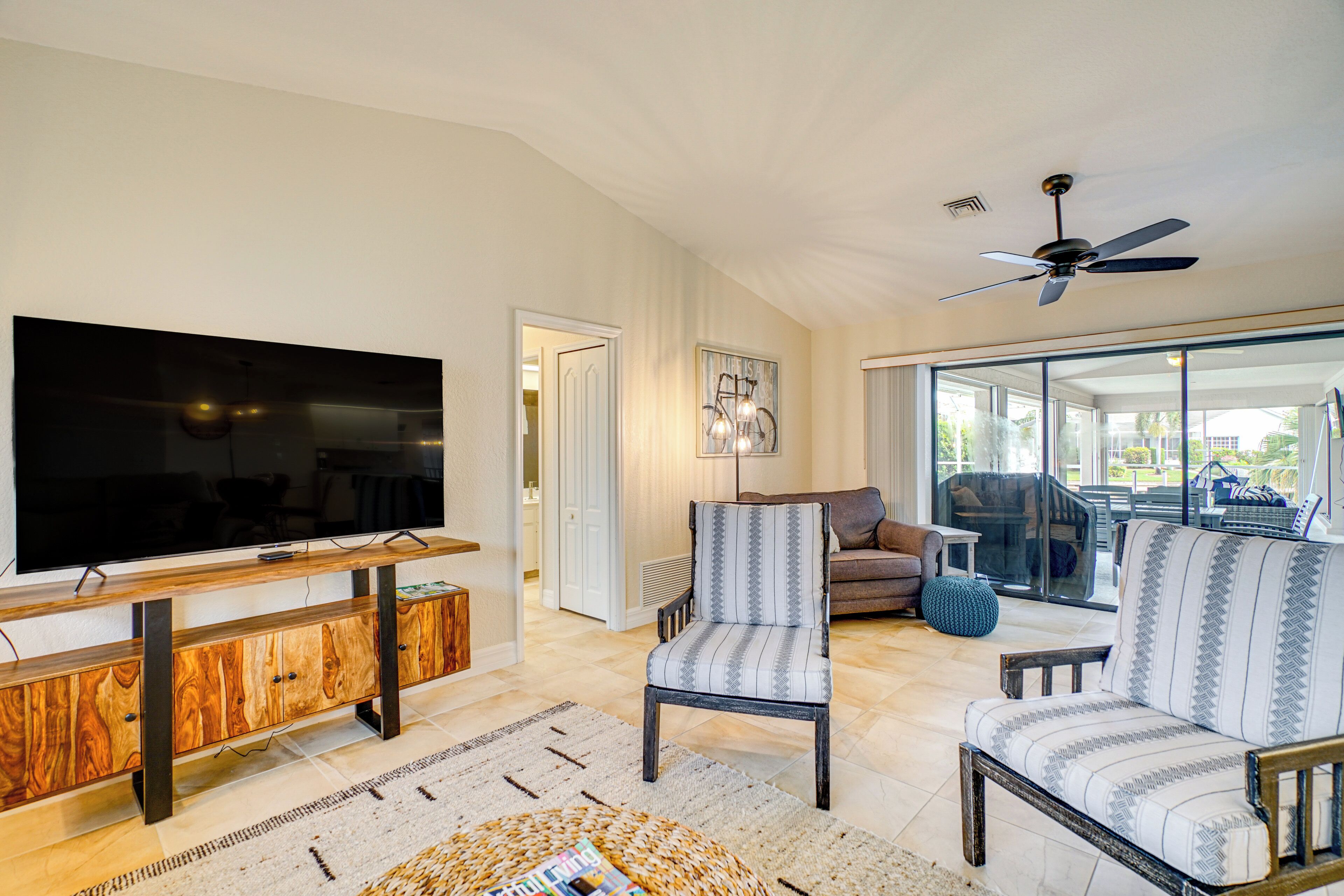A living room with a flat-screen TV, a ceiling fan, and a large window.