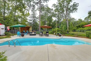 A swimming pool with a concrete deck, surrounded by trees and a covered seating area with chairs and umbrellas. - Backyard Oasis w/ Pool: Sunny Sumter Studio! (Sumter)