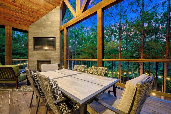 An outdoor deck with a dining table and chairs, surrounded by wooden beams and large windows overlooking a forest. - Broken Bow Home w/ Hot Tub, Game Room & EV Charger (Broken Bow)