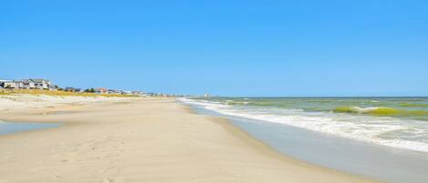 A wide sandy beach with gentle waves, a clear blue sky, and houses in the background.