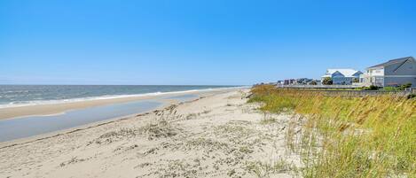 A beach with a sandy shore, grassy dunes, and a row of houses along the water's edge.
