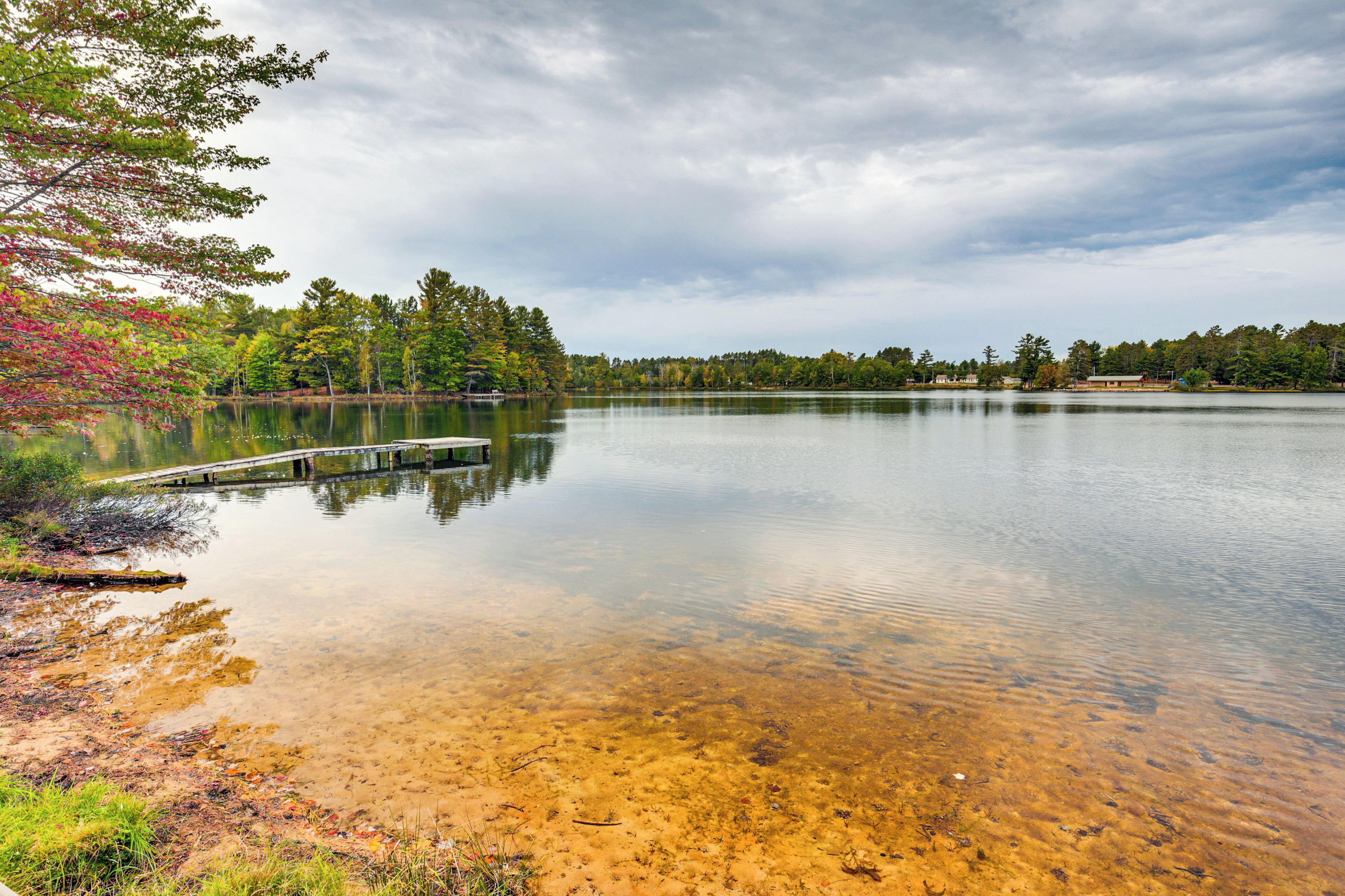A calm lake with a wooden dock extending into the water, surrounded by trees and a cloudy sky.