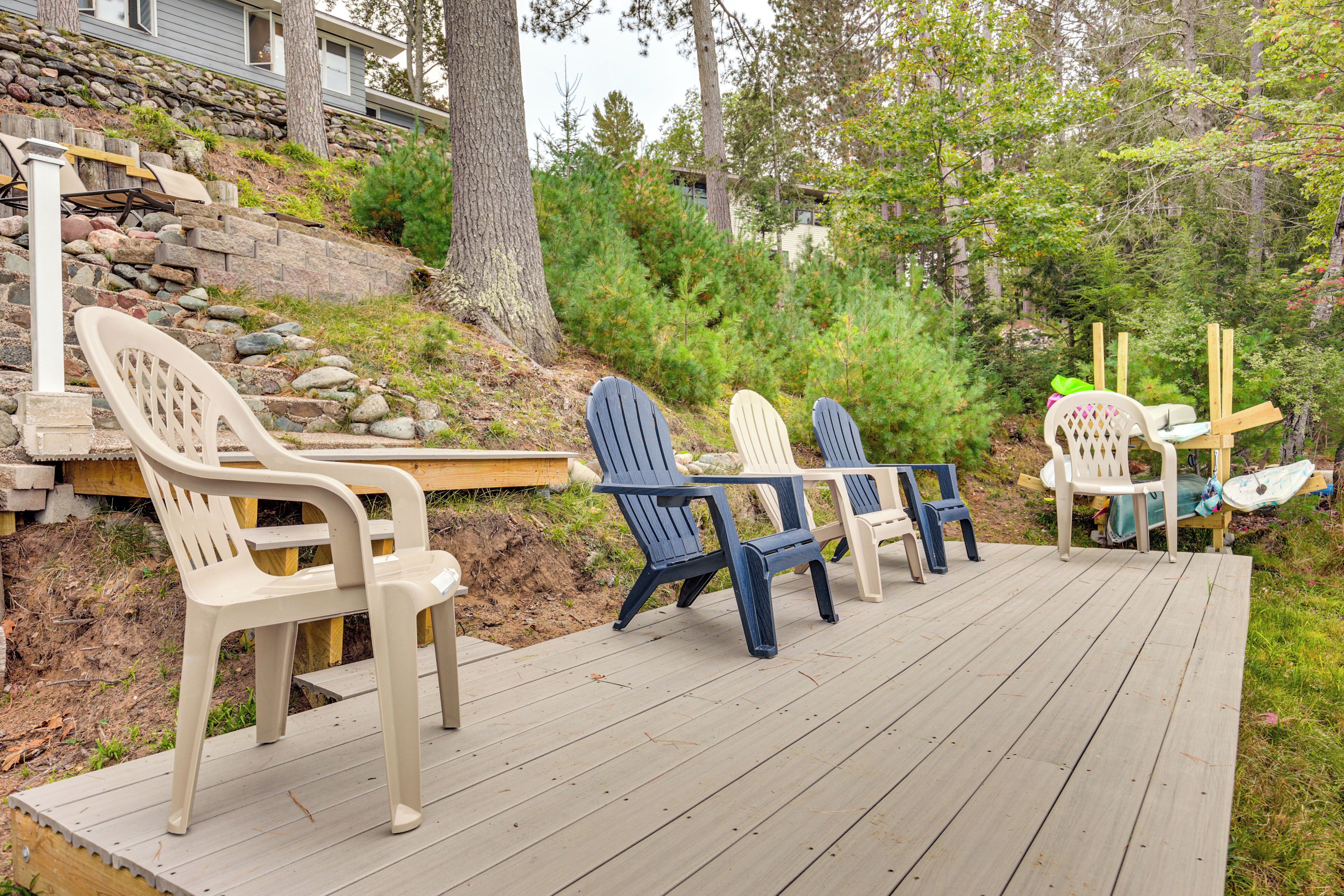 A wooden deck with Adirondack chairs and a table, surrounded by trees and a stone retaining wall.
