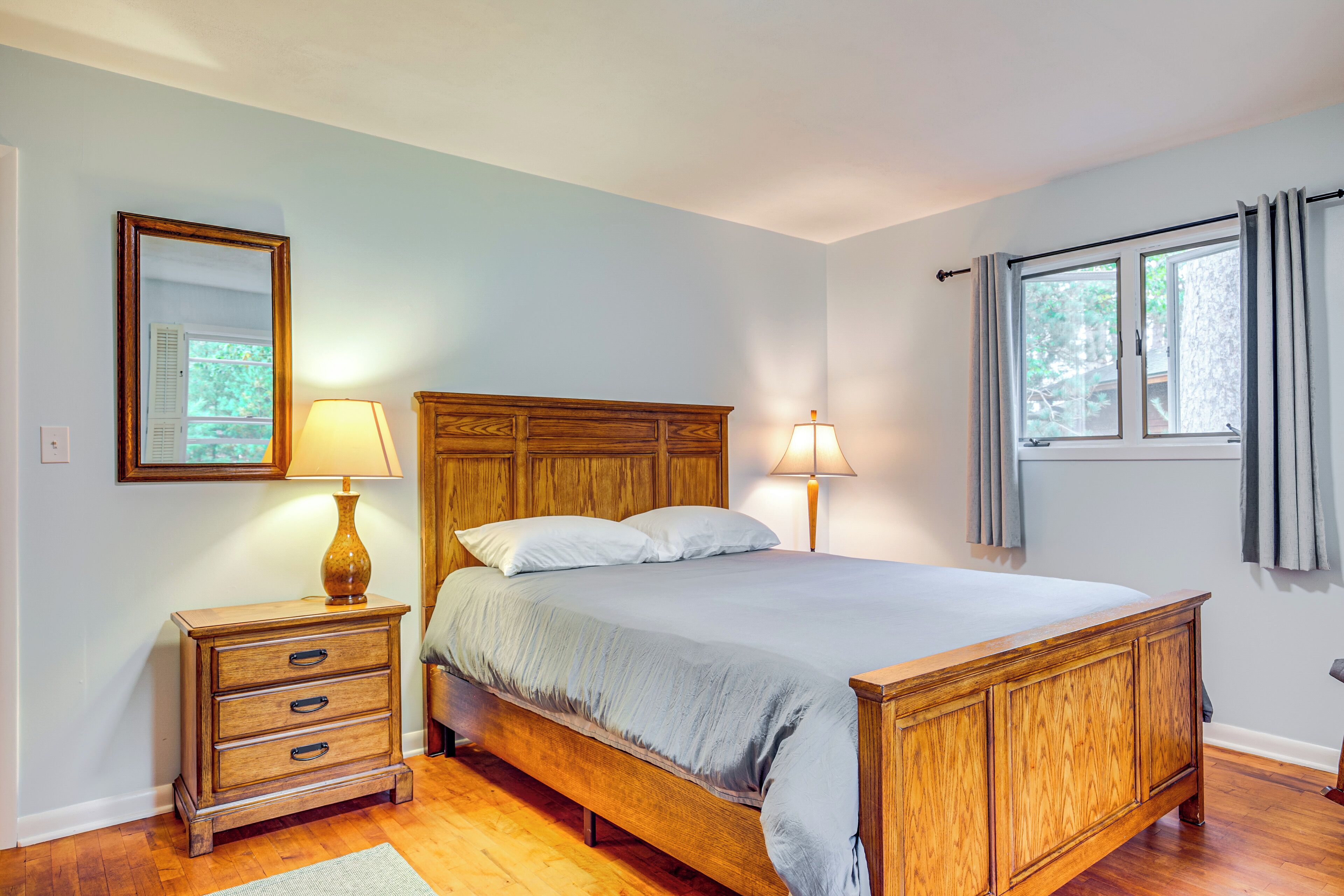 Quarto com cama de madeira, duas mesas de cabeceira com luminárias, um espelho e uma janela com cortinas.