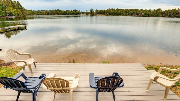 Eine Holzterrasse mit vier Stühlen und einem Tisch, mit Blick auf einen ruhigen See, umgeben von Bäumen.