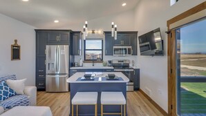 A modern kitchen with dark cabinets, stainless steel appliances, a central island with stools, and a large window offering a view. - "montana Blue" w/ Grill Near Skiing! (Kalispell)