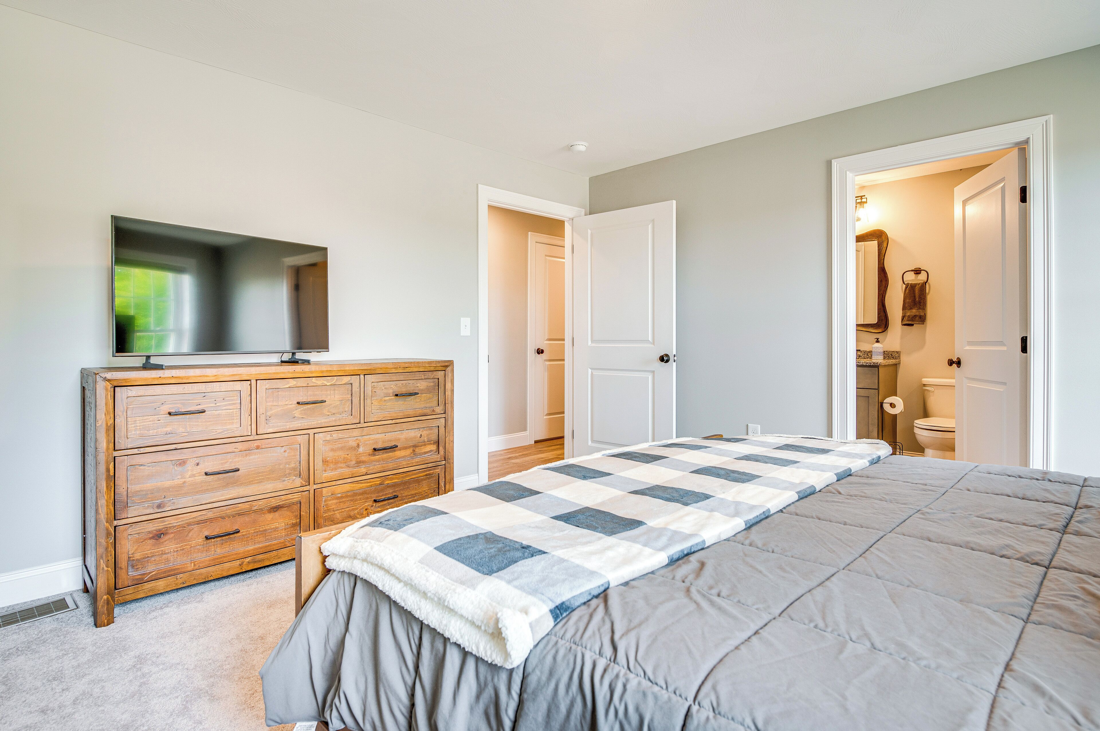 A bedroom with a large bed, a wooden dresser, a television, and a bathroom visible through an open door.