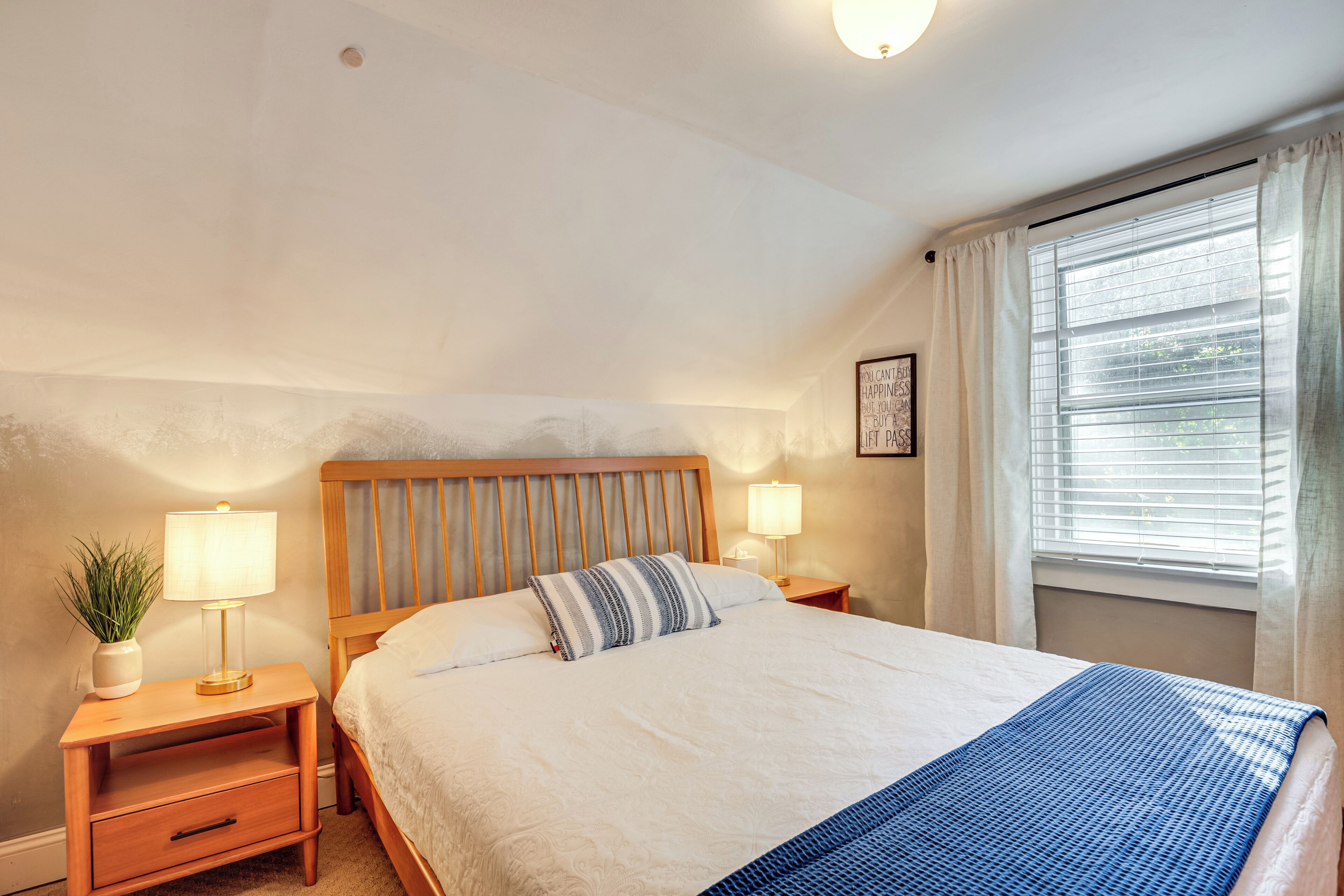 A neatly made bed with a wooden headboard, two bedside tables with lamps, a window with blinds, and a small plant on the left side.