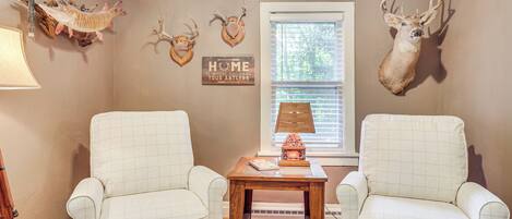 A room with two white armchairs, a wooden table with a lamp, and wall-mounted deer antlers.