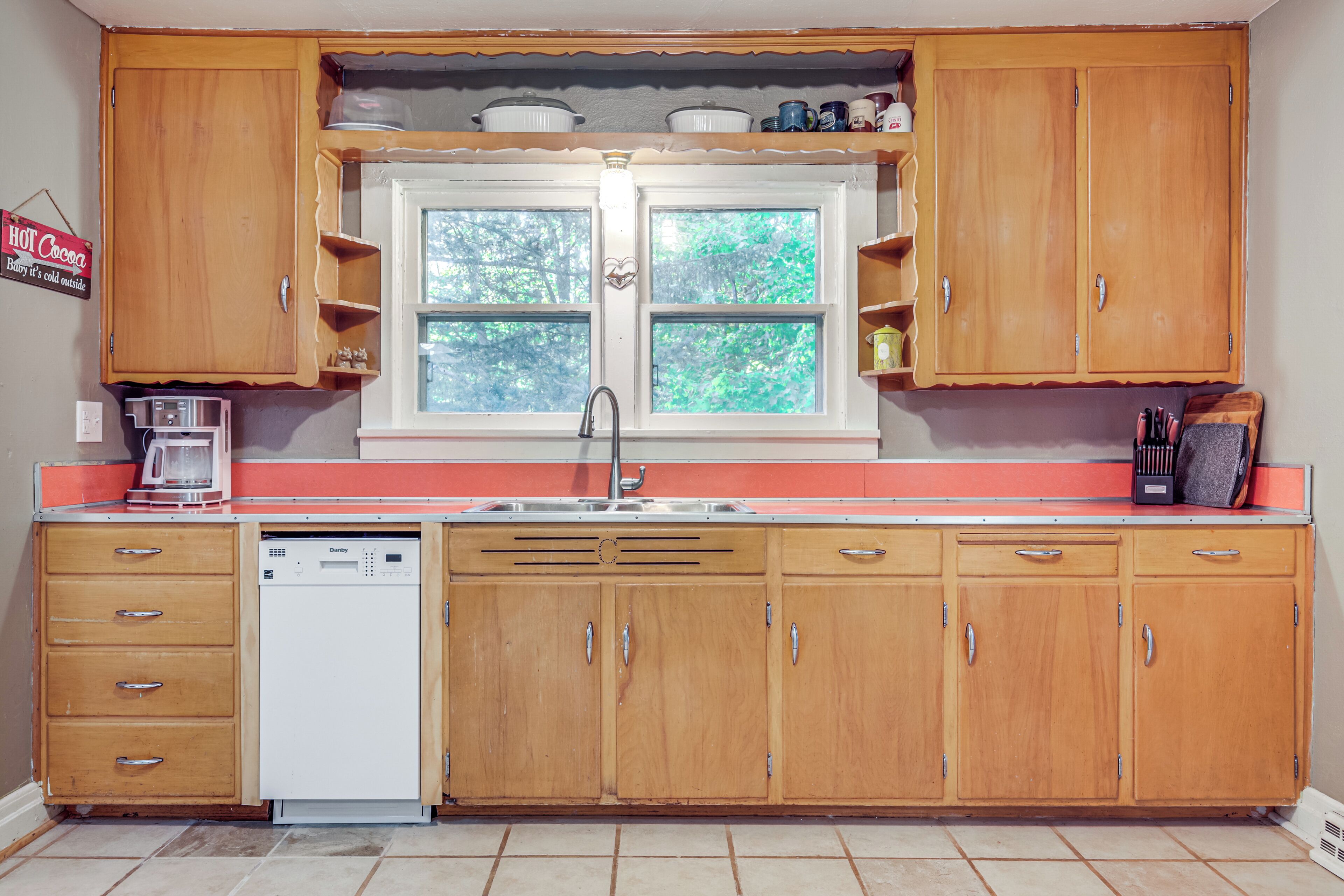 A kitchen with wooden cabinets, a dishwasher, and a window with a view of trees.