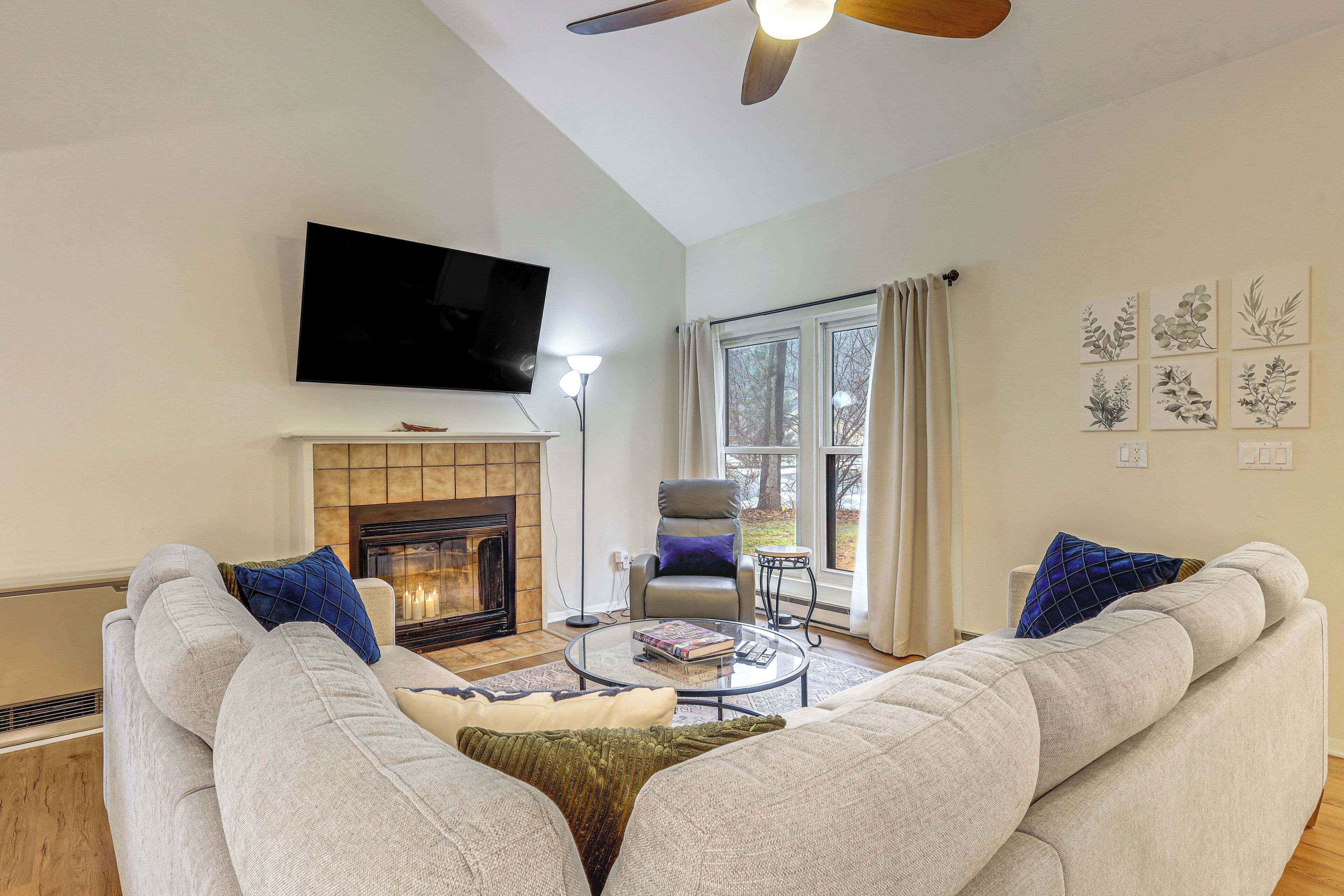 A living room with a fireplace, a flat-screen TV, a ceiling fan, and a large window with curtains.