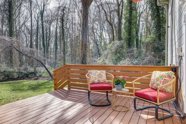 Une terrasse en bois, agrémentée de fauteuils à bascule en osier et d’une petite table, le tout entouré d’une zone boisée.