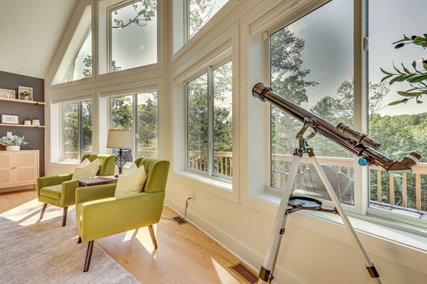 Una habitación con ventanas amplias, un telescopio, sillones verdes, suelo de madera y un armario blanco.