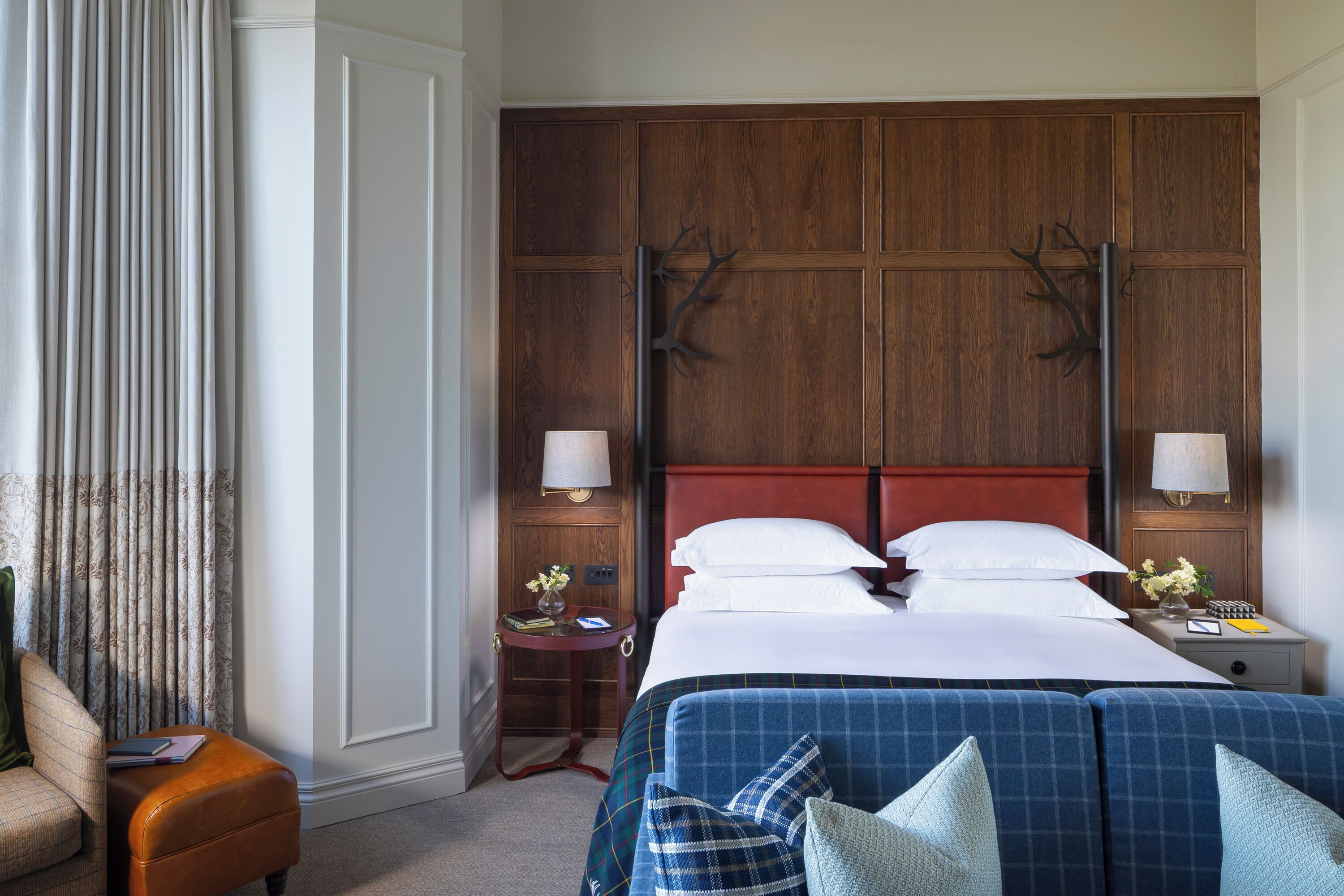 Une chambre d’hôtel avec une tête de lit en bois, un lit avec du linge de lit blanc, un canapé bleu, une petite table avec une lampe et une chaise.
