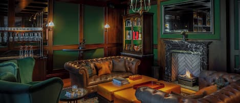 A cozy lounge with leather sofas, a fireplace, a bar area with glasses, and a green wall.