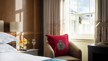 Hotellihuone, jossa on sänky, punaisella koristetyynyllä varustettu tuoli, yöpöytä lampun kanssa, maljakko kukkineen ja ikkuna, jossa on verhot.