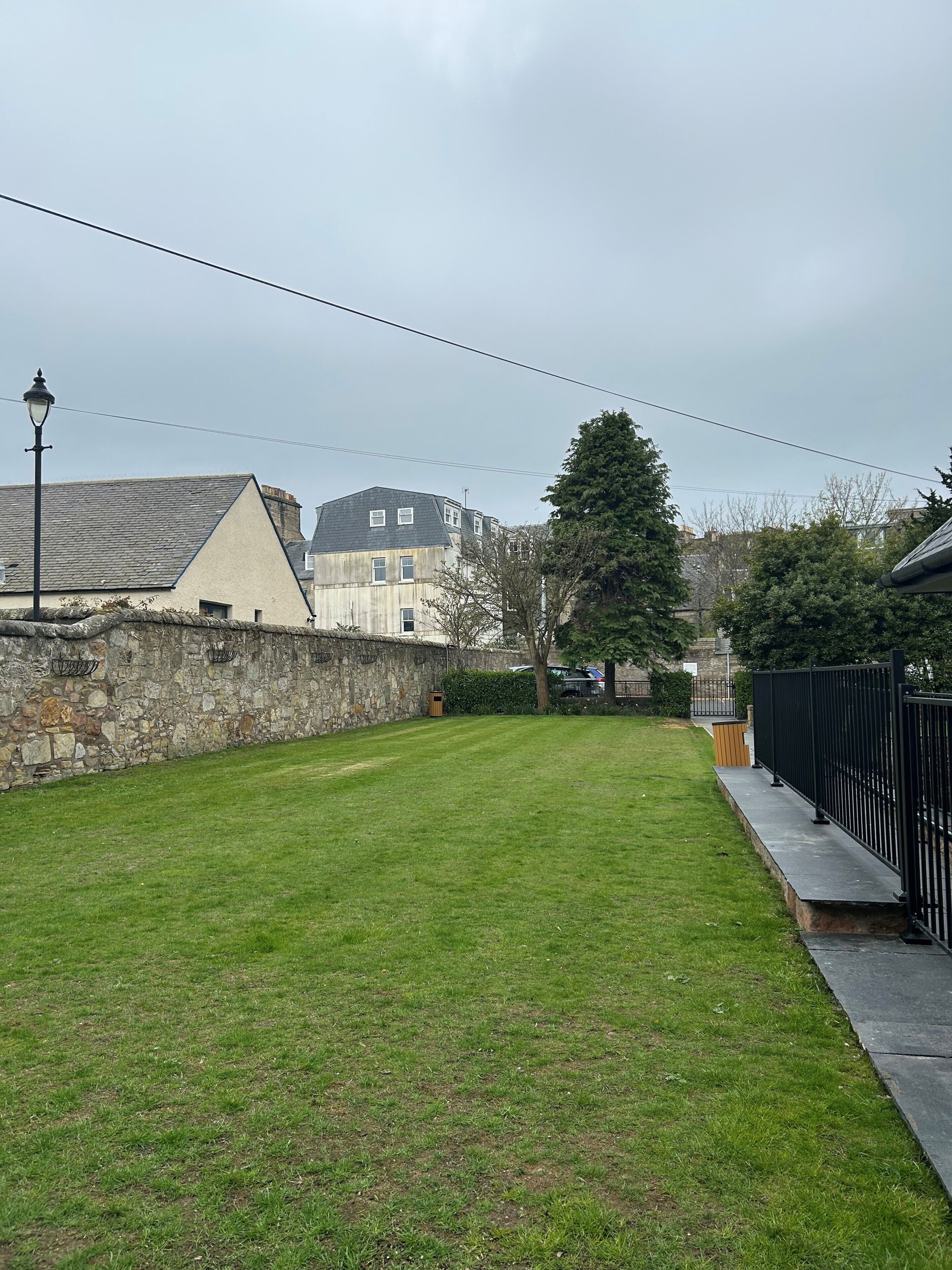 A grassy backyard with a stone wall, a lamp post, and a black metal fence.