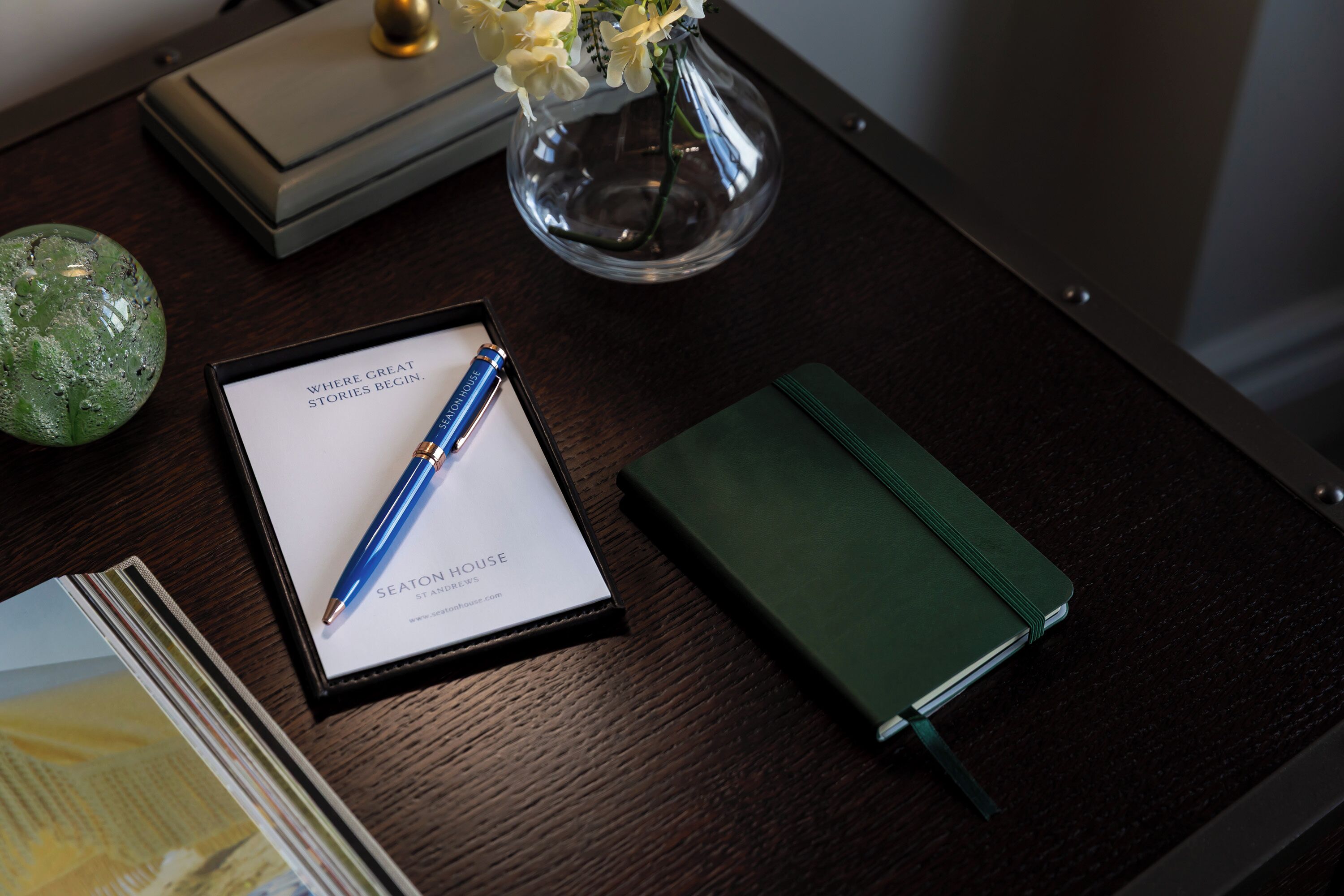 Un escritorio con una libreta, un bolígrafo, un florero de vidrio con flores y un objeto decorativo.