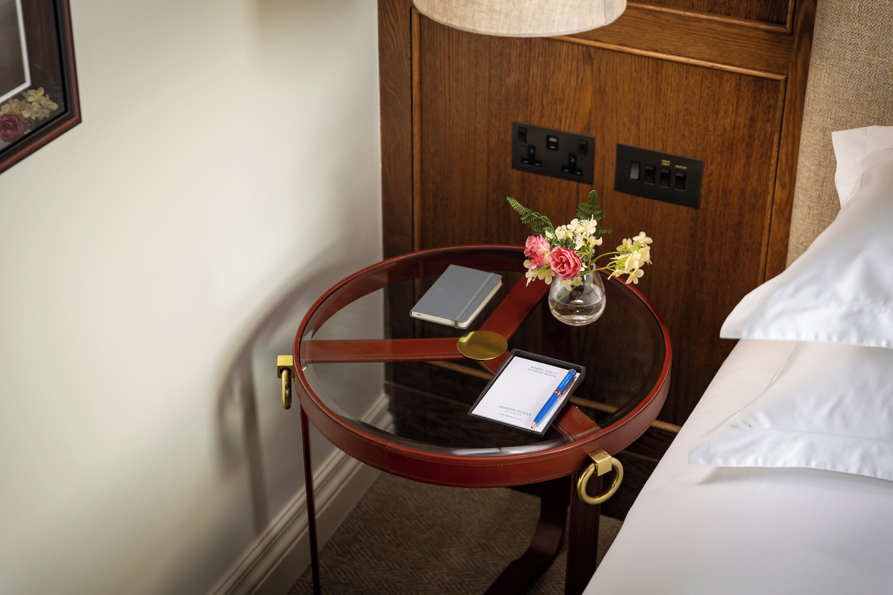 Habitación de hotel con una mesa redonda de vidrio, un florero con flores, un teléfono inteligente y una tableta.