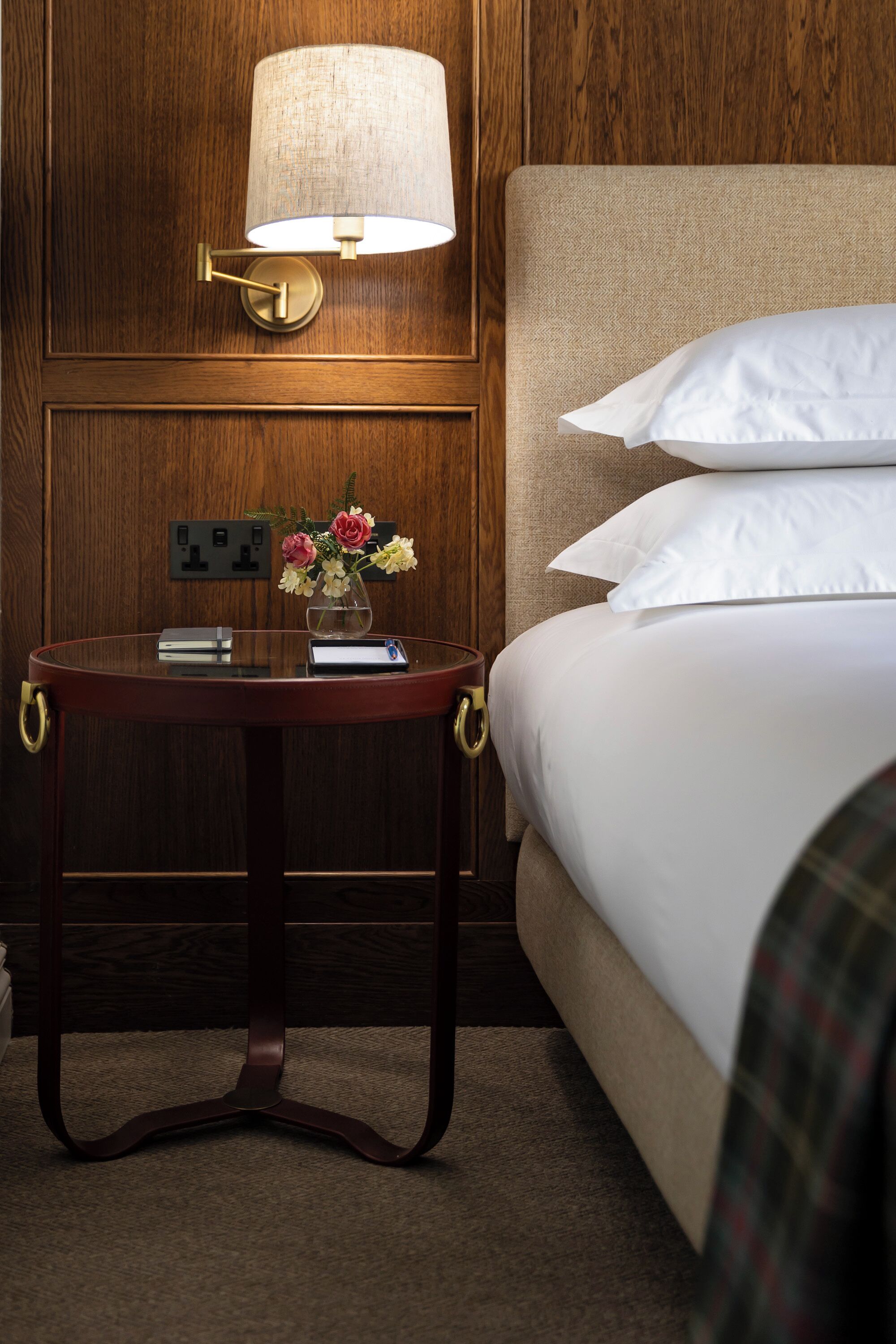 A hotel room with a wooden panel wall, a round table with a lamp, a vase with flowers, and a bed with white linens.