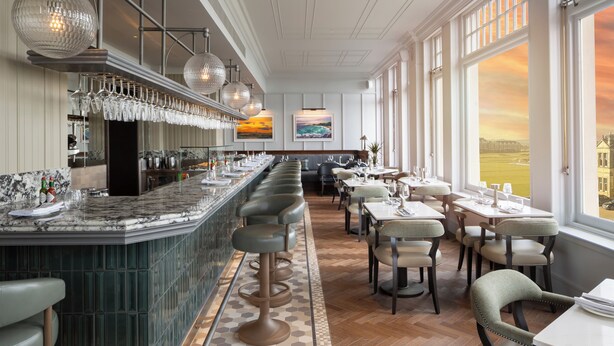 Een modern restaurant met een lange bar, bekleedde barkrukken en uitzicht op de ondergaande zon door grote ramen.