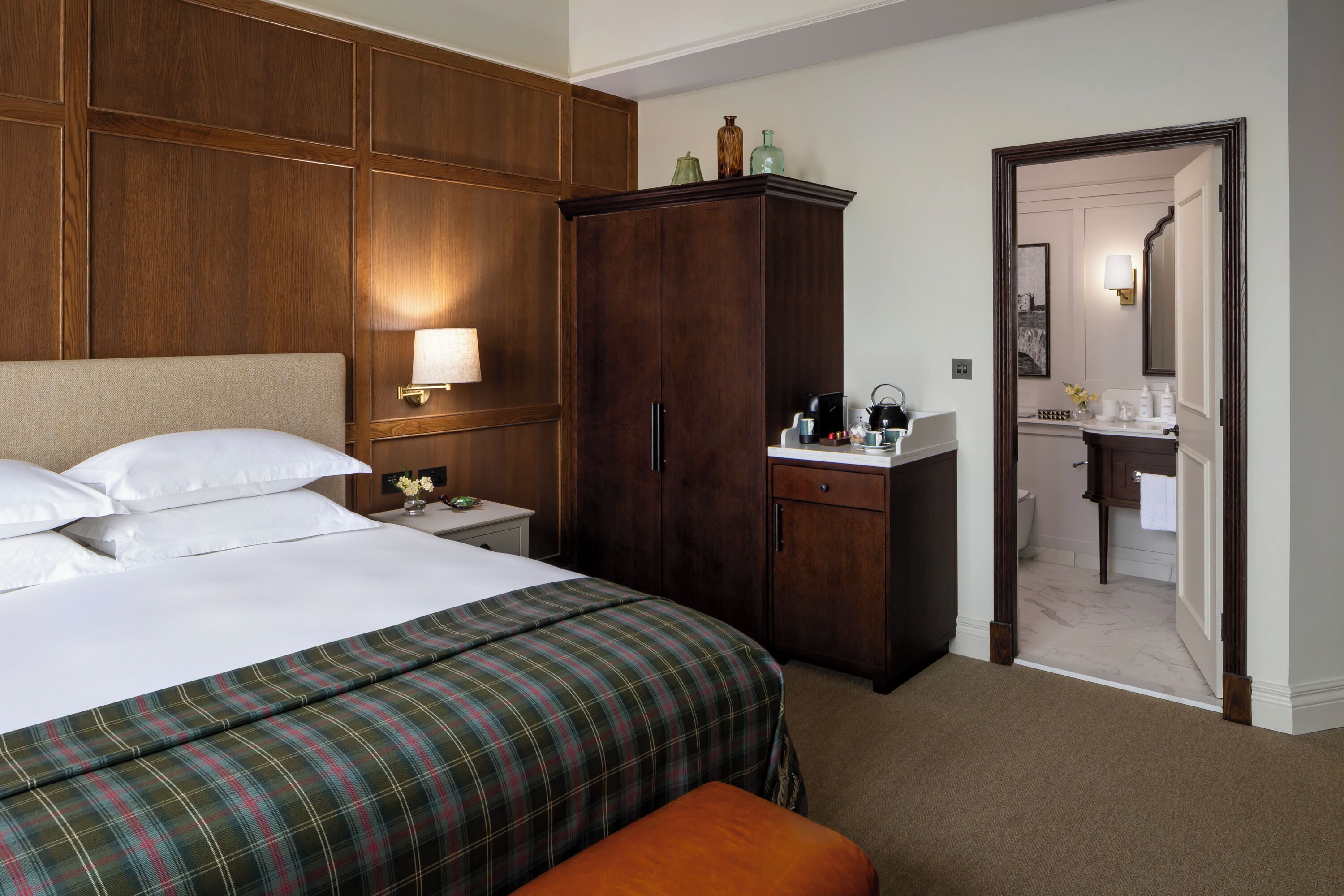 A hotel room with a large bed, a wooden wardrobe, a nightstand with a lamp, and a bathroom visible through an open door.
