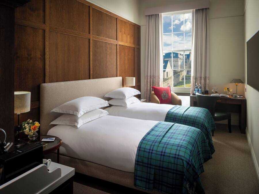 Habitación de hotel con dos camas, un escritorio, una silla, un televisor y una ventana con vistas a un castillo.