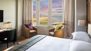 Een hotelkamer met een groot bed, twee fauteuils, een bureau met lamp en uitzicht op het strand door een groot raam.
