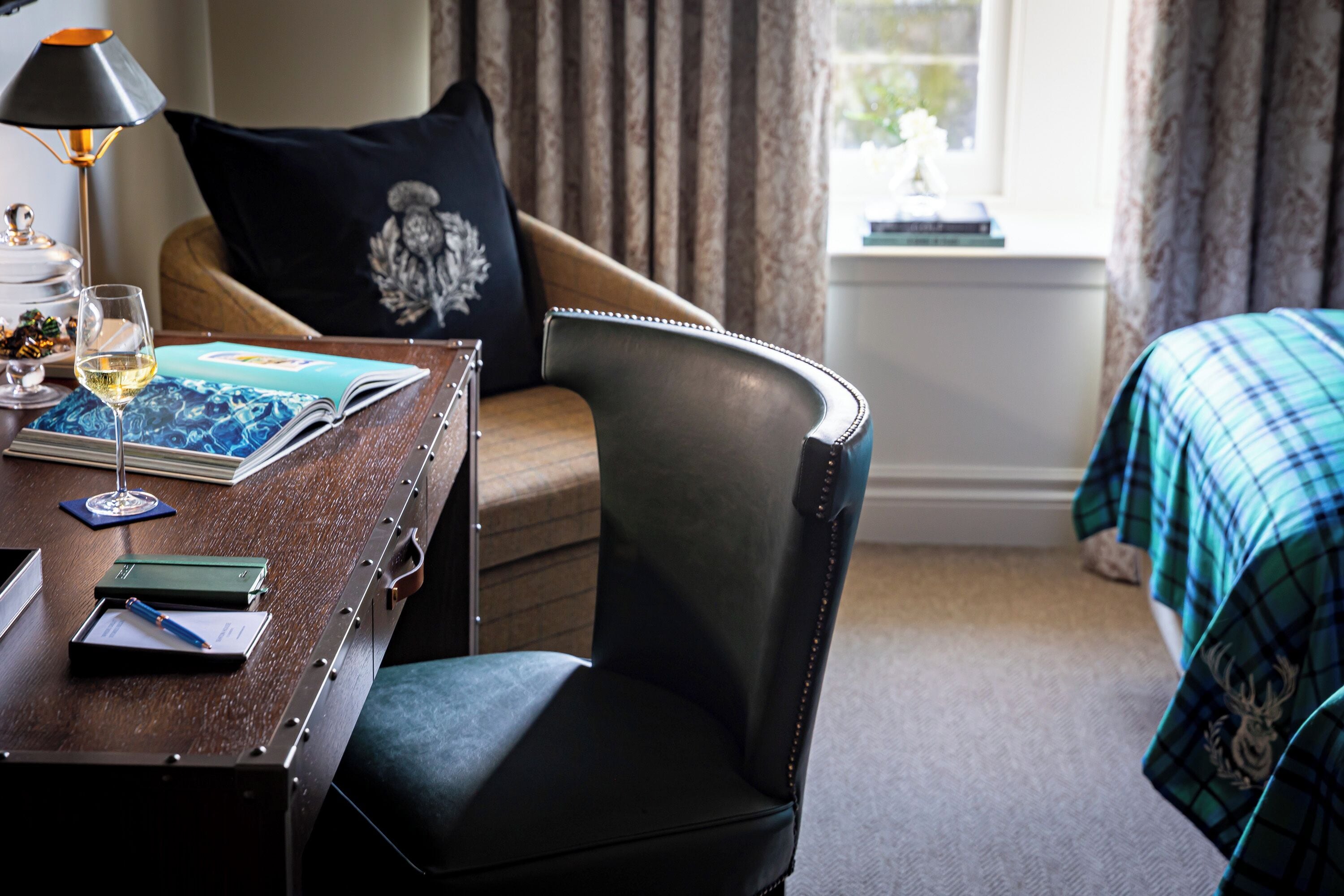 A hotel room with a wooden desk, a leather chair, a bed with a plaid blanket, a window with curtains, and a lamp.
