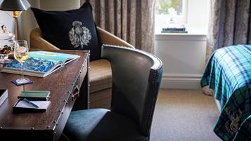 A hotel room with a wooden desk, a leather chair, a bed with a plaid blanket, a window with curtains, and a lamp.